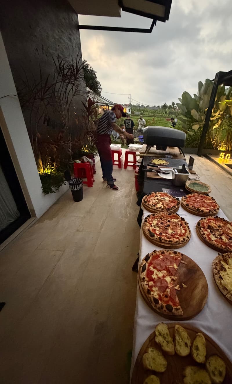 A long table of pizzas beside the oven with rice fields behind, in Ubud