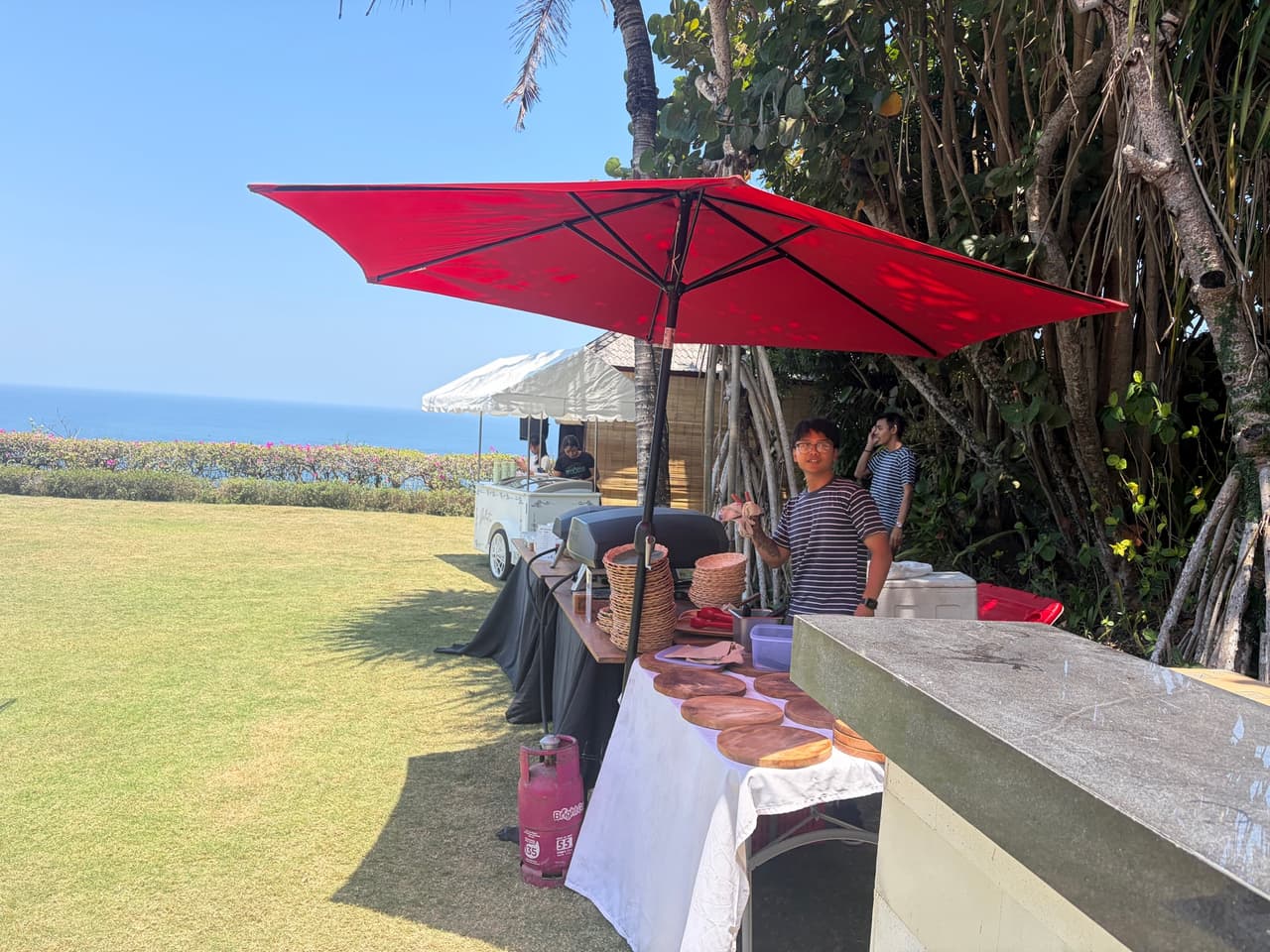 Our pizza station under a red umbrella on a clifftop lawn in Uluwatu, with the ocean behind